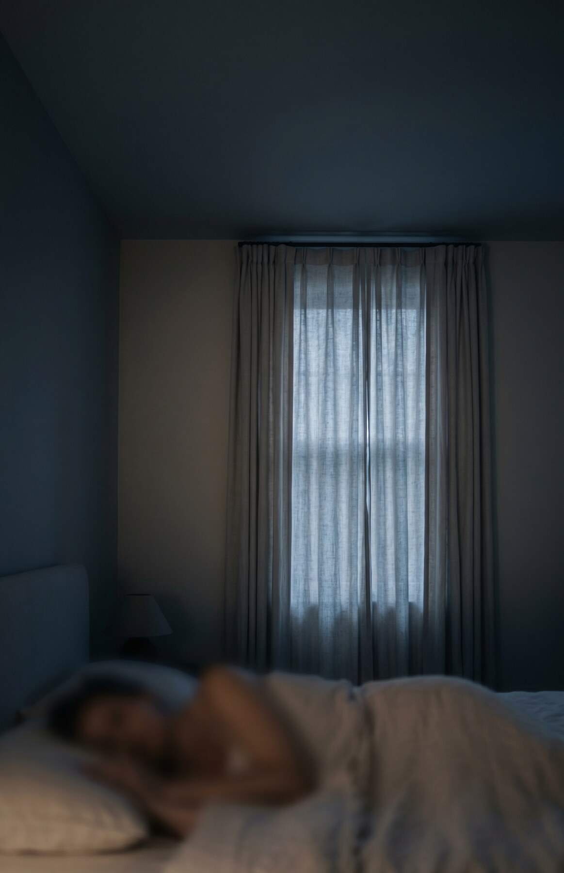 A dimly lit bedroom in the middle of the night, with cool light filtering through linen curtains onto the foot of a made bed.